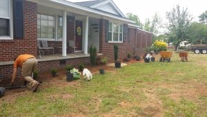 Landscaper installs shrubs in front of red brick house with porch, surrounded by tools, soil bags, and wheelbarrows during setup.
