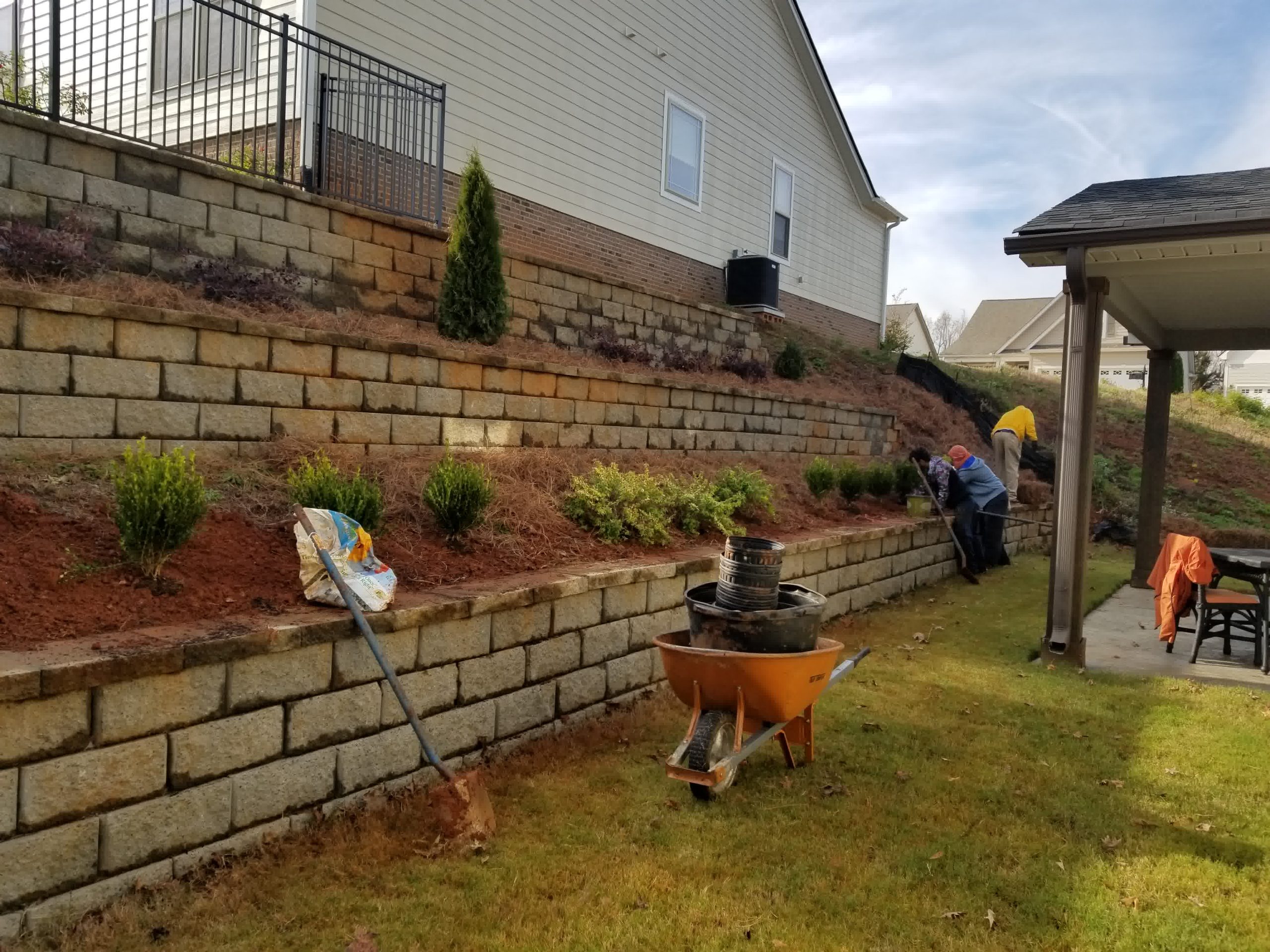 Workers install shrubs along a tiered retaining wall with stacked stone blocks on a sloped backyard hill.



