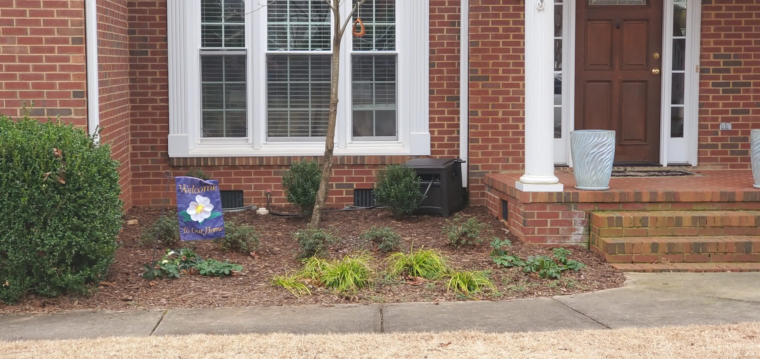 Front porch flower bed with mulch, small evergreen bushes, a welcome flag, and a young tree centered under large front windows.
