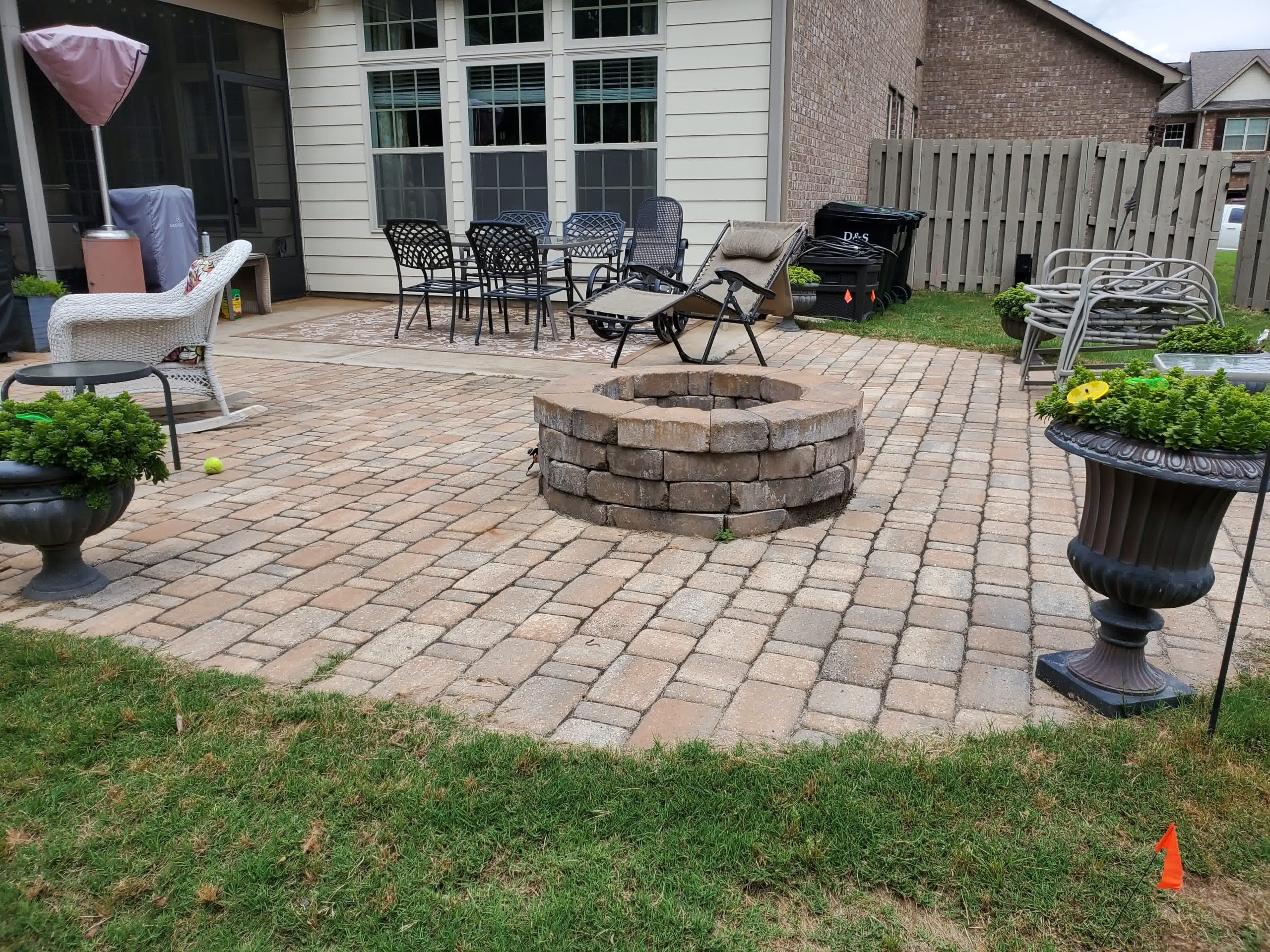 Backyard patio with interlocking stone pavers, central fire pit, lounge furniture, and potted greenery.

