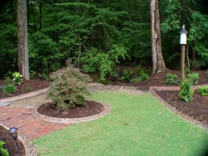 Lush backyard landscape with brick paver path, manicured lawn, ornamental tree, and dense green foliage near a forest creek.