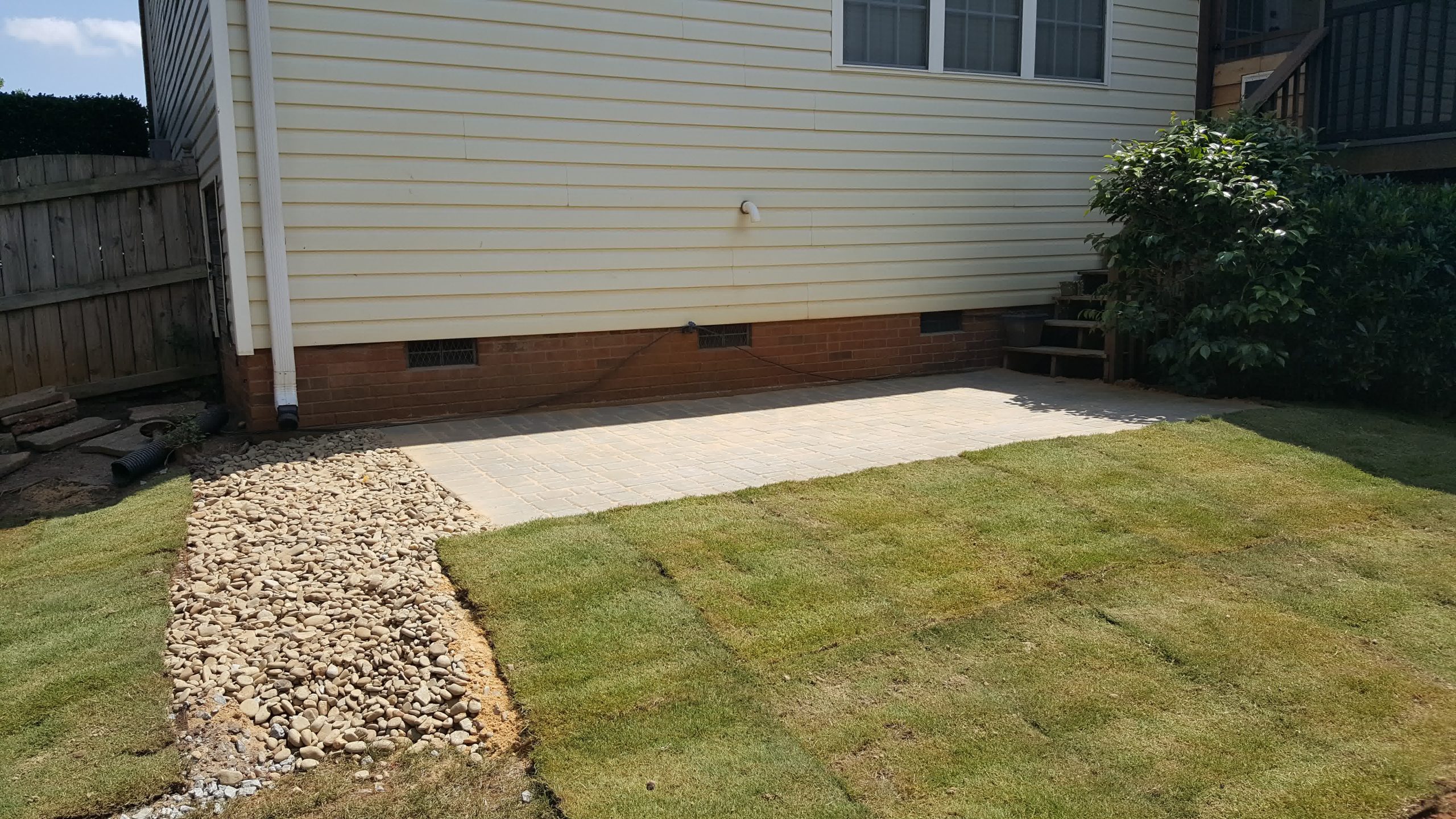 solomons sod installation inman sc scaled - Solomon's Landscape & Design Fresh sod installed alongside a stone drainage path and paver patio at the back of a home with beige siding and brick foundation.